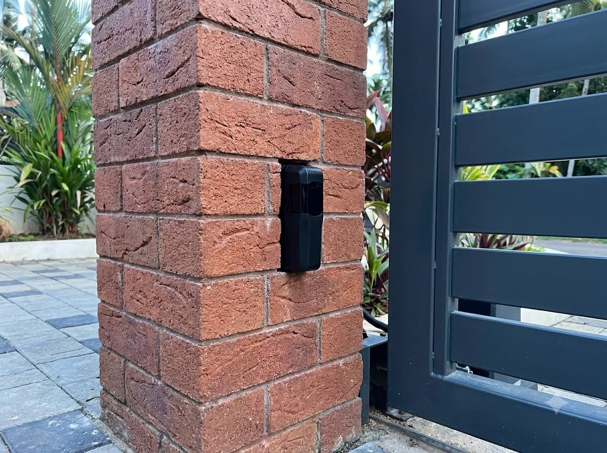 Photocell safety sensor mounted on a brick gate pillar beside a dark metal gate